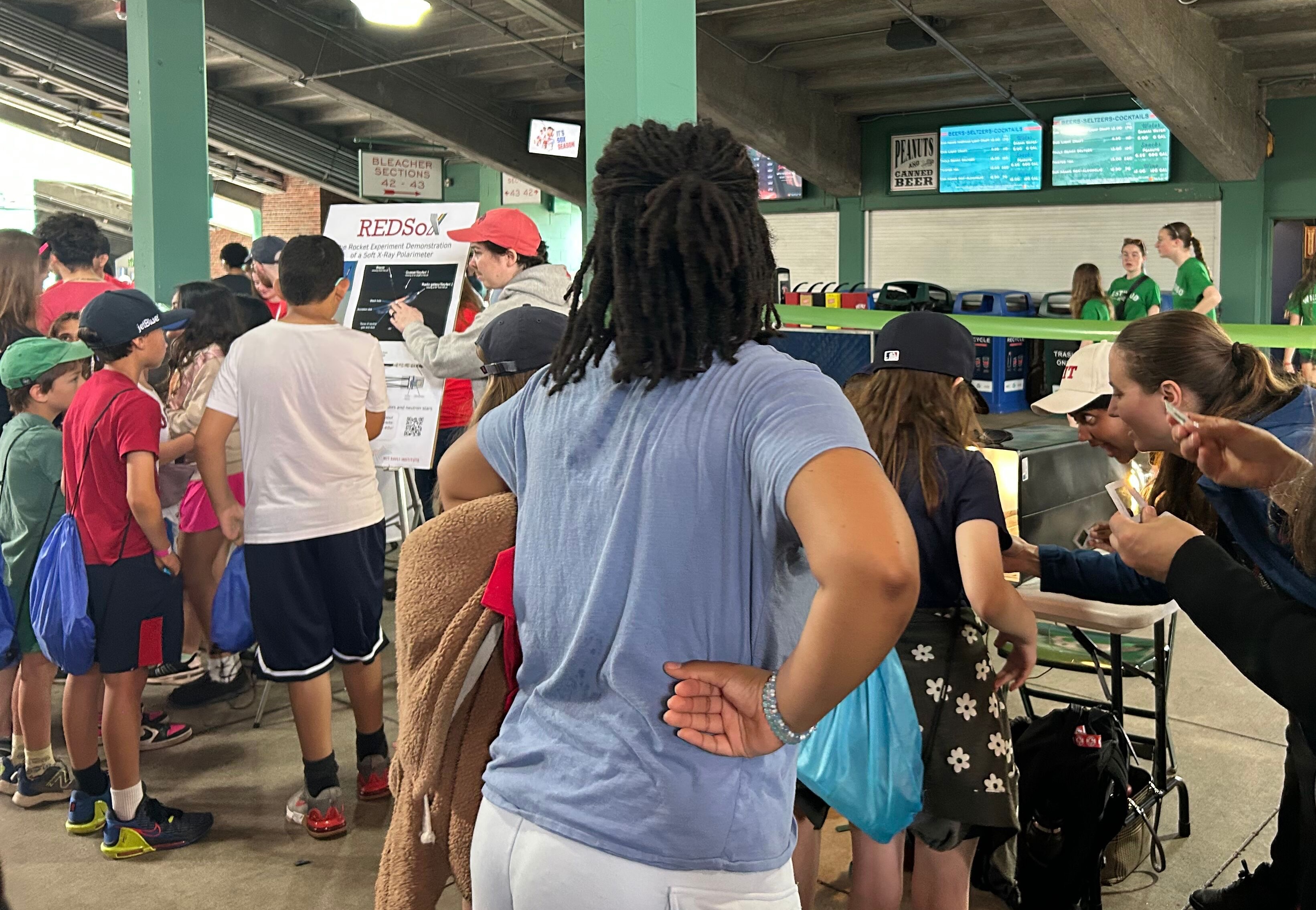 A group of students learns about the REDSoX rocket project from MIT staff, one explaining a poster, two demonstrating polarized light.