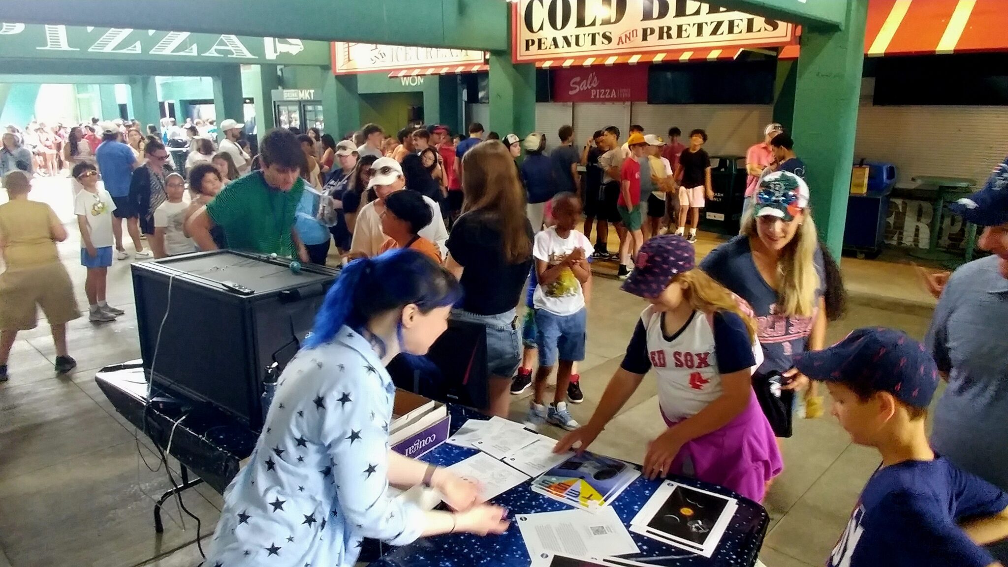 A woman at a table providing paper handouts about exoplanets to people wearing Red Sox gear.