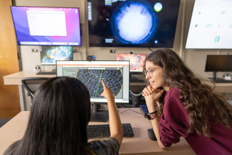 Students Erin Cusson and Niko Kopparapu confer in front of an array of monitors with astronomy data.