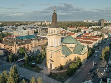 erial view of a yellow church with a green dome and tall steeple in the center of a town, surrounded by buildings and trees.