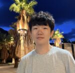 A photo of Te wearing a grey shirt in front of palm trees at night.