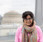 Nergis stands wearing a scarf, pink top, and glasses with the MIT Dome in view behind her.