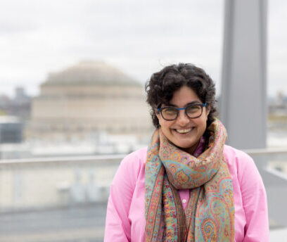 Nergis stands wearing a scarf, pink top, and glasses with the MIT Dome in view behind her.