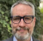 A photo of Scott wearing glasses with foliage behind.