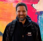 Nicholas wears a black MIT Physics jacket with colorful background behind.