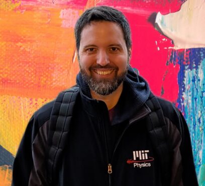 Nicholas wears a black MIT Physics jacket with colorful background behind.