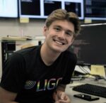 Matthew smiles, wearing a LIGO shirt in front of a computer work station.