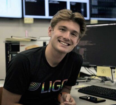 Matthew smiles, wearing a LIGO shirt in front of a computer work station.