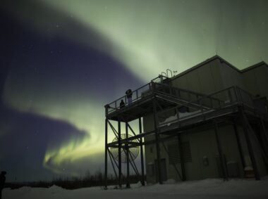 Two students observe aurora borealis arcs above observation facilities at Poker Flat Research Range.