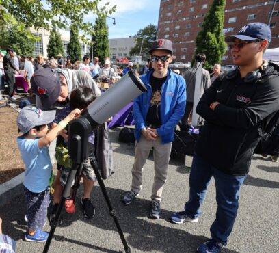 a child looks through a large telescope in daytime while instructors look on.