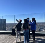 a group of people stands on a roof deck at twilight, looking up at the sky with a telescope next to them