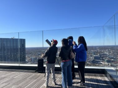 a group of people stands on a roof deck at twilight, looking up at the sky with a telescope next to them