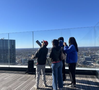 a group of people stands on a roof deck at twilight, looking up at the sky with a telescope next to them
